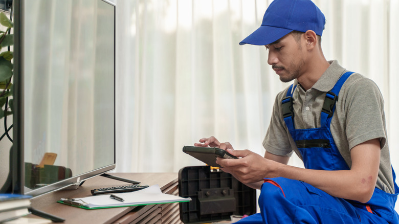 Man in jumpsuit with hat nails down in front of flat screen TV