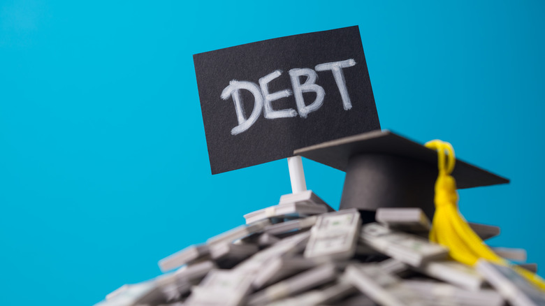 Small card that says debt sits beside small graduation cap on pile of tiny stacks of money