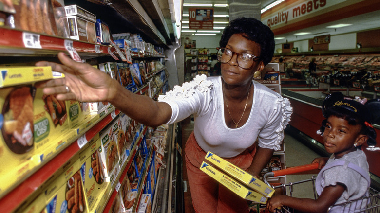 Woman with child in grocery cart holds packaged food as she reaches for another food item