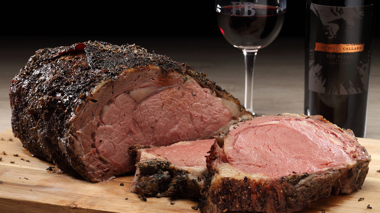 Closeup of a thick cut of prime rib on a wooden cutting board in front of a glass and bottle of red wine.