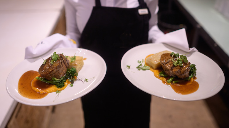 A server at a steakhouse carrying two plates of thick-cut steaks
