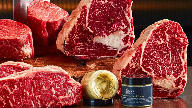 Multiple cuts of steak on a butcher counter with jars of seasonings and herb butter.