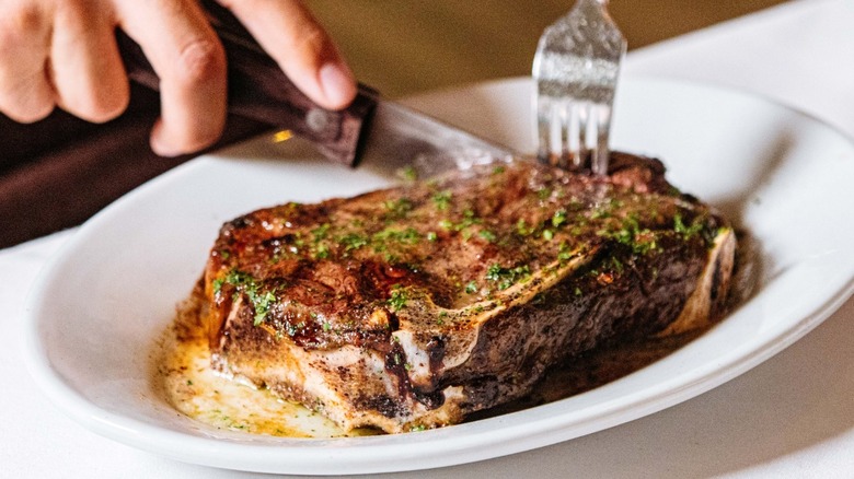 A diner cutting a buttery steak garnished with herbs on a white platter at Ruth's Chris Steak House.