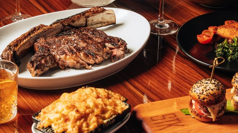 An off-the-bone steak on a white platter beside sliders and macaroni and cheese on a polished wooden table.