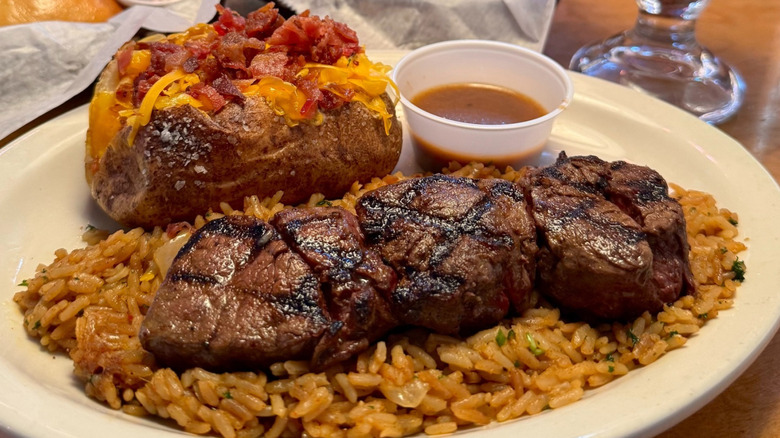 A close-up of medallion steaks on a bed of rice beside a loaded baked potato with a side of gravy on a white plate at Texas Roadhouse.