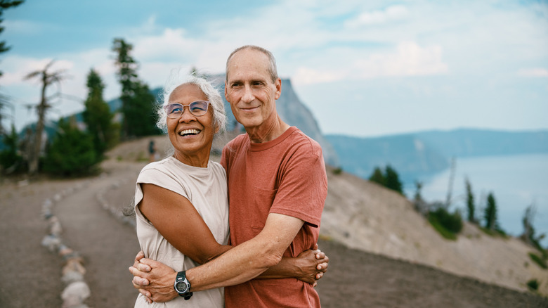 A Pacific Islander senior woman and her Caucasian husband who have recently retired embrace and smile while enjoying the view of Crater Lake during nature hike.