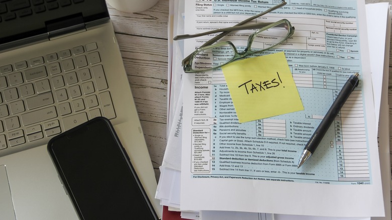 Laptop, coffee mug, smartphone, glasses, pen, and tax forms with a sticky note that says "taxes!"