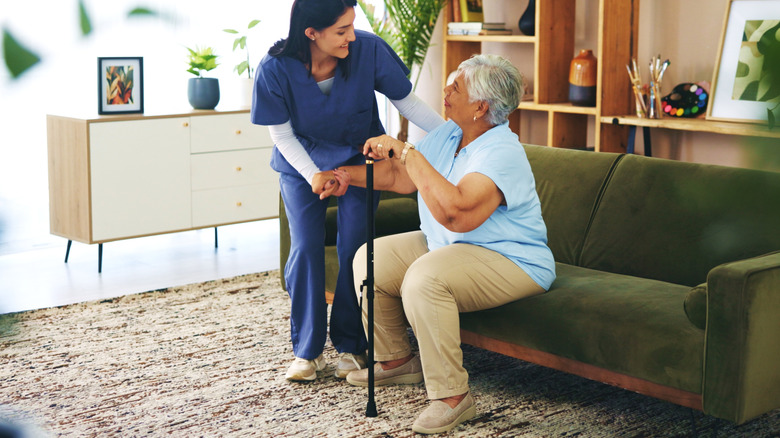 Woman in nurse uniform helps mature woman holding cane and sitting on couch