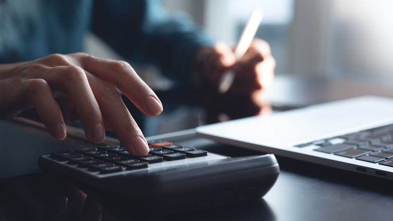 fingers on a calculator near a laptop while the other hand holds a pen