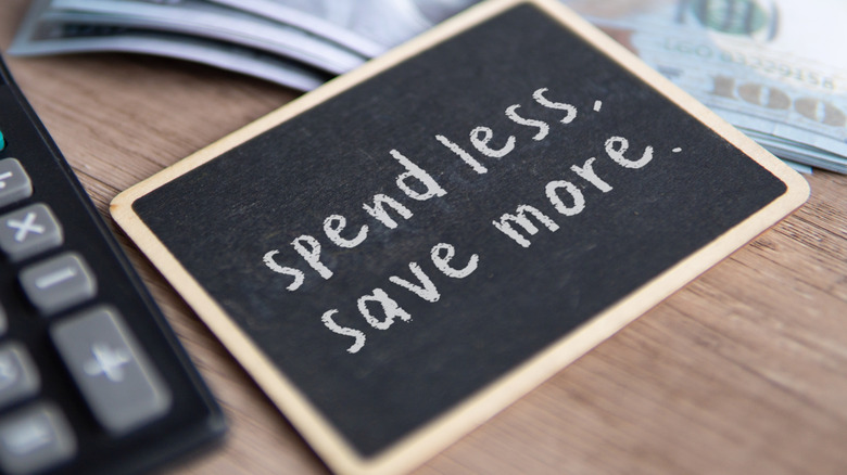 chalkboard with "spend less, save more" written, with calculator and stack of $100 bills