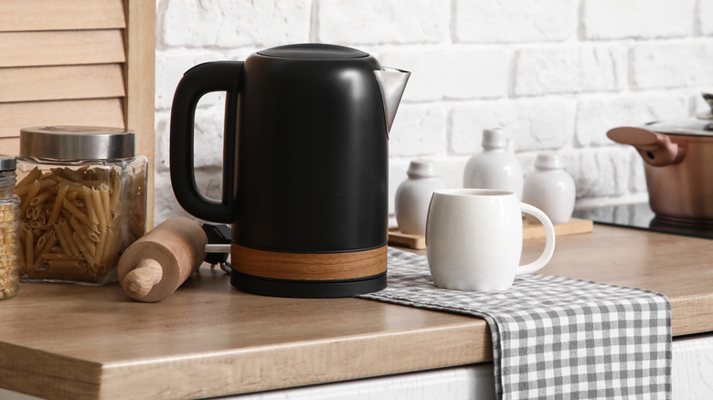 An electric kettle rest on a table top near a container with pasta, a rolling pin, and a small mug