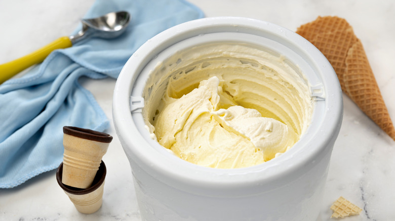 Top view of an ice cream maker with vanilla ice cream inside, surrounded by a cloth, ice cream scoop, and cones