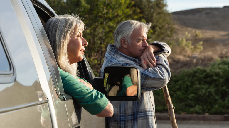 An old woman looks out of a pickup truck window while her husband stands beside her