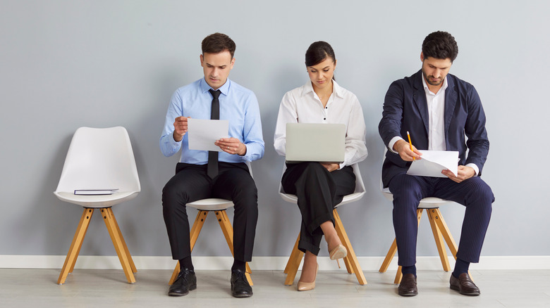 Men holding documents and a woman with a laptop sit in chairs against a wall.