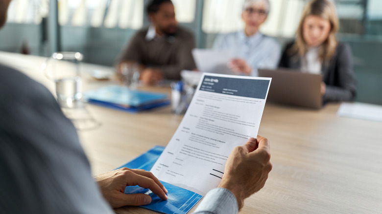 A man looks over a resume at table with a blurred group of office workers seated across from him.
