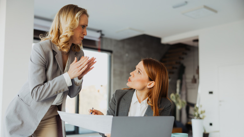 Standing woman listens to seated woman with open laptop holding a holding document and a pen.