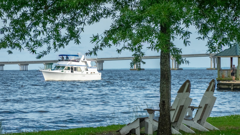 Boat on the water with bridge in the background, tree and wooden lawn chairs in foreground