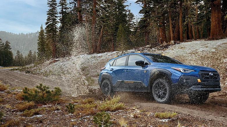 blue 2026 Subaru Crosstrek splashing through mud puddle on road through tree-covered mountainous area