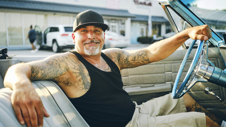 An aging hipster with tattoos smiling in the driver's seat of a convertible car with the roof down.