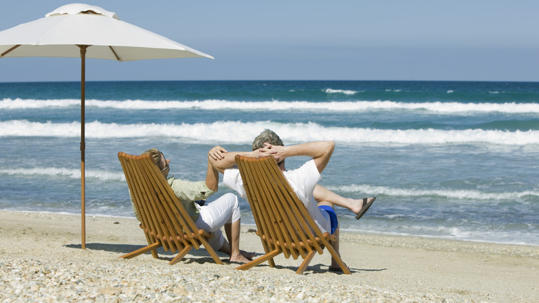 A mature couple sitting on bamboo beach chairs while holding hands on a beach.