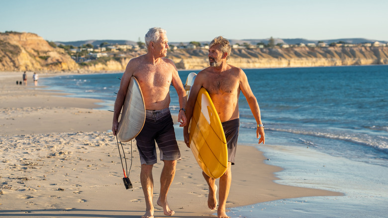 Two mature men carrying surfboards while walking on a beach.
