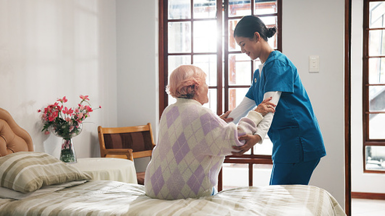 A smiling nurse helping an elderly woman out of bed in a nursing home.