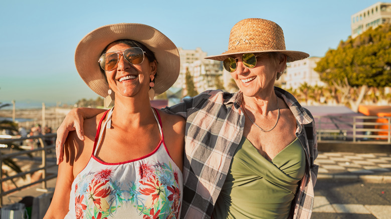 Two smiling mature woman wearing sunglasses on a beachside boardwalk on a sunny day.