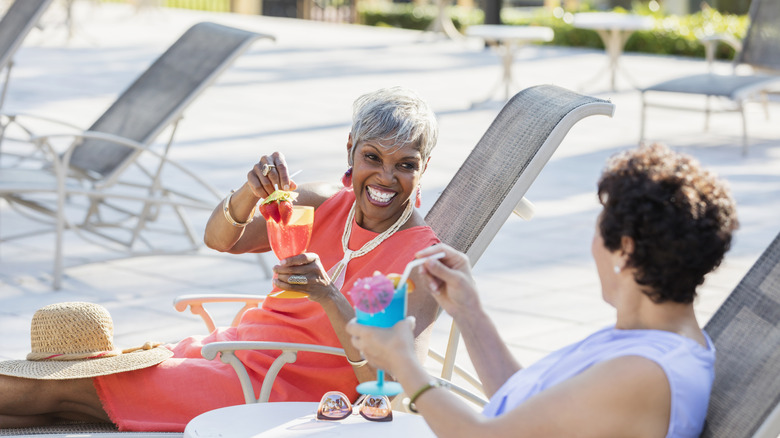 Two distinguished mature women laughing while holding cocktails in pool chairs on a sunny day.