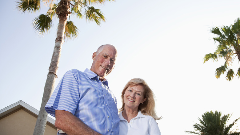 A smiling senior couple standing in front of a house and palm trees in Florida.