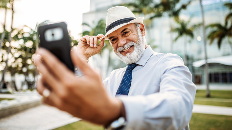 A well dressed mature man taking a selfie with faded palm trees in the background.
