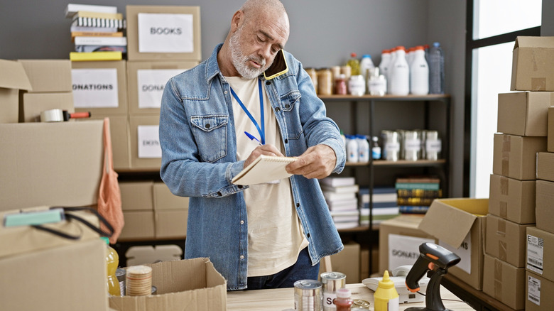 A middle-aged man writing on a notepad in a storage room surrounded by cardboard boxes and shelving.