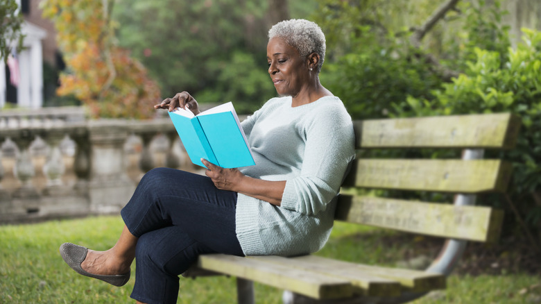 A mature woman sitting on a park bench while reading a book.