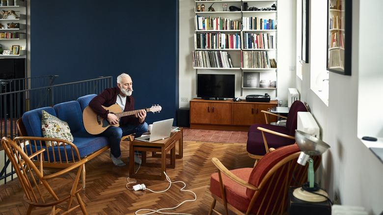 A senior man sitting on a sofa playing guitar while surrounded by furniture.