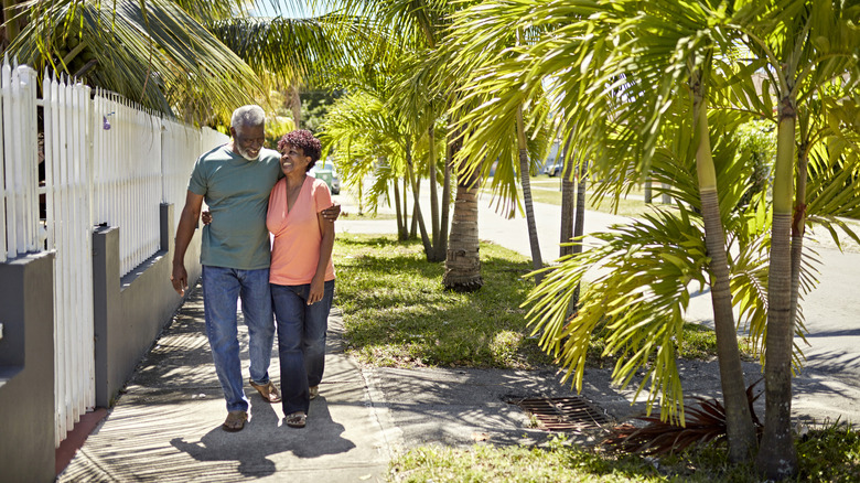 Full length view of Black couple in their 70s wearing casual clothing and approaching camera with arms around each other, talking, and laughing.