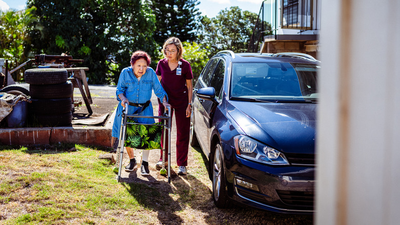 A female nurse using a strap to help an elderly woman cross the driveway on a sunny day.