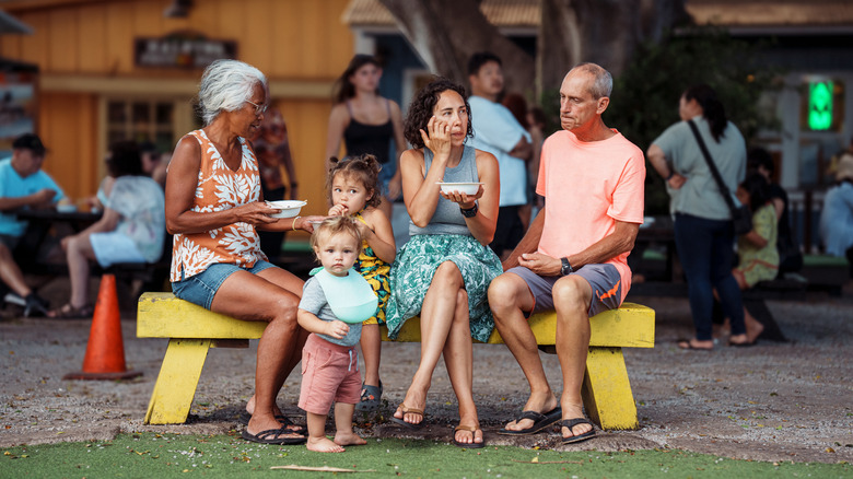 A happy multi-generation family sits on a bench together at an outdoor market in Hawaii.