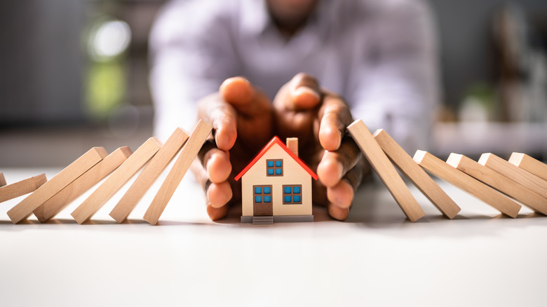 Man holding hands over small model home blocking small wooden dominos from falling onto model home.