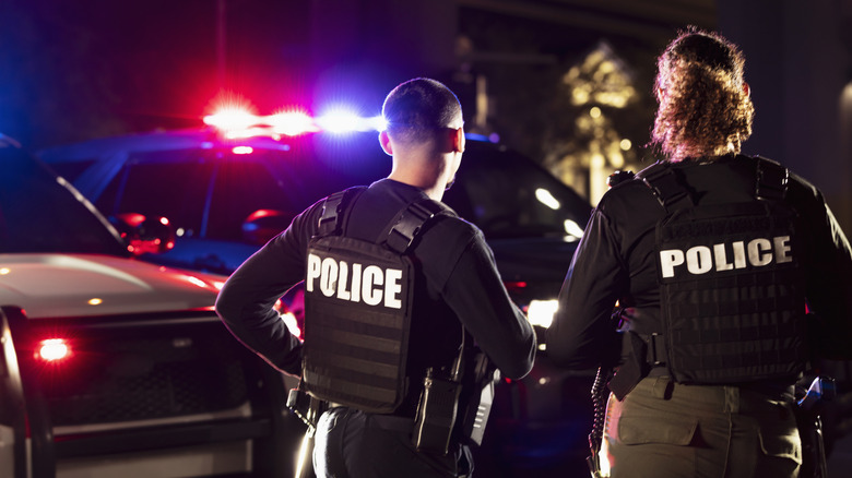 Rear view of two multiracial police officers standing side by side in front of their police vehicles with emergency lights on.