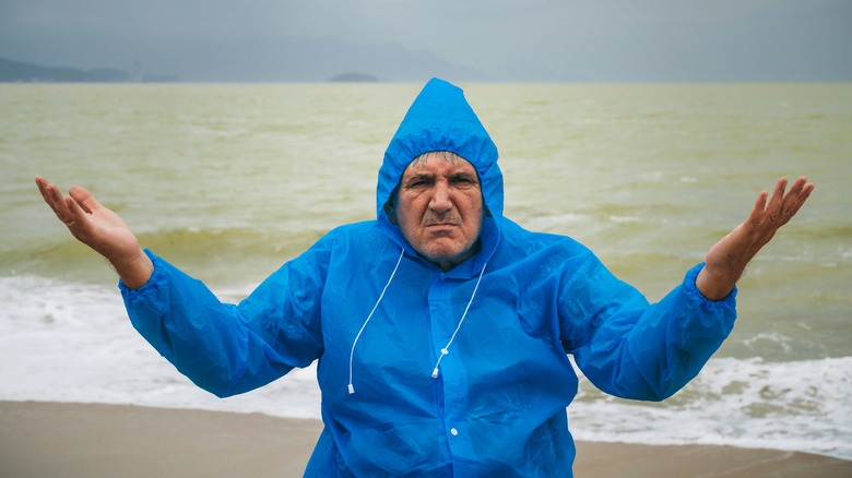 Frowning older man in rain coat stands in front of rough beach waves