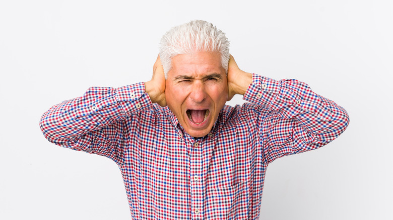 Older man in long sleeve dress shirt covers ears while yelling