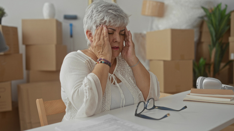 Senior grey haired plus size woman removes glasses and rubs temples amid stacked moving boxes in building.