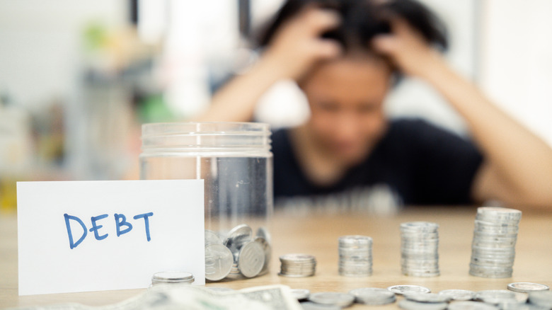 A woman sits at a table, looking distressed, with piled coins and a debt note.