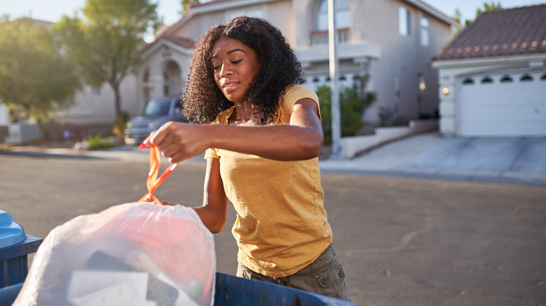 A young woman putting a trashbag in a bin on a suburban street