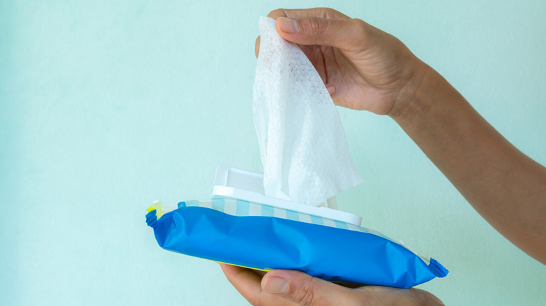A hand pulling a wet wipe up and out of a plastic package against a light blue backdrop