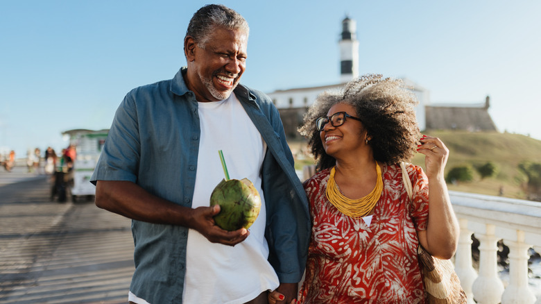 Happy older couple walking on boardwalk while the man holds a coconut drink
