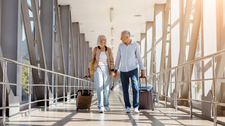 A retired couple holding hands in a long sunlit hallway while wheeling carry-on luggage behind them.