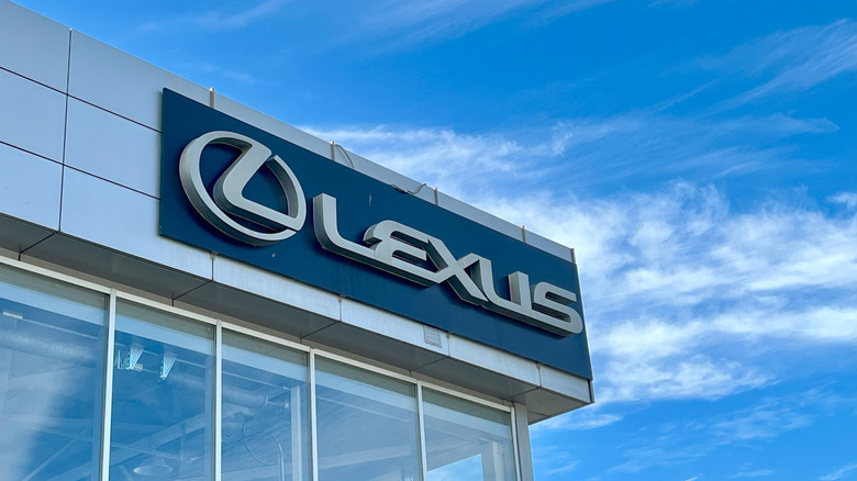 Lexus brand logo on dealership building with clouds in background.