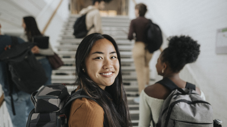 A smiling young woman looking at the camera as college students ascend stairs in the background.