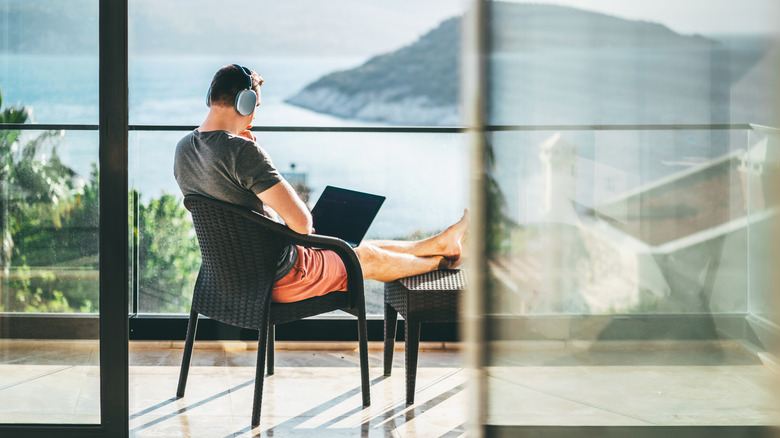 A man wearing headphones sitting on a balcony with a laptop in his lap.