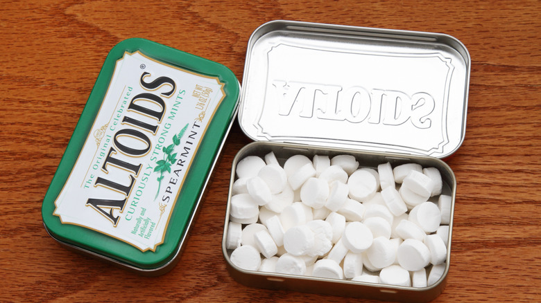 A closed green and white tin of Altoids next to an open tin container with dozens of small white mint candies